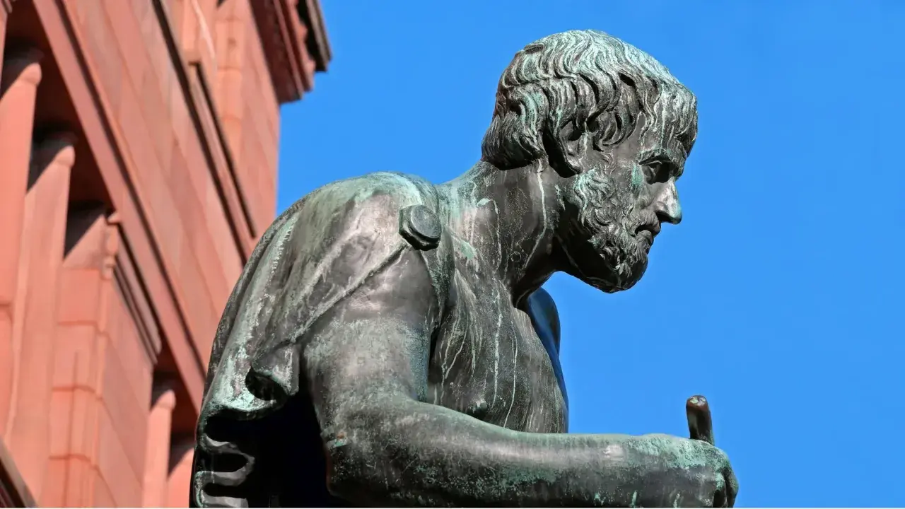 Estatua de bronce de Aristóteles, pensador cuyas frases inspiran la sabiduría. El cielo azul y un edificio de ladrillo rojo completan la escena.