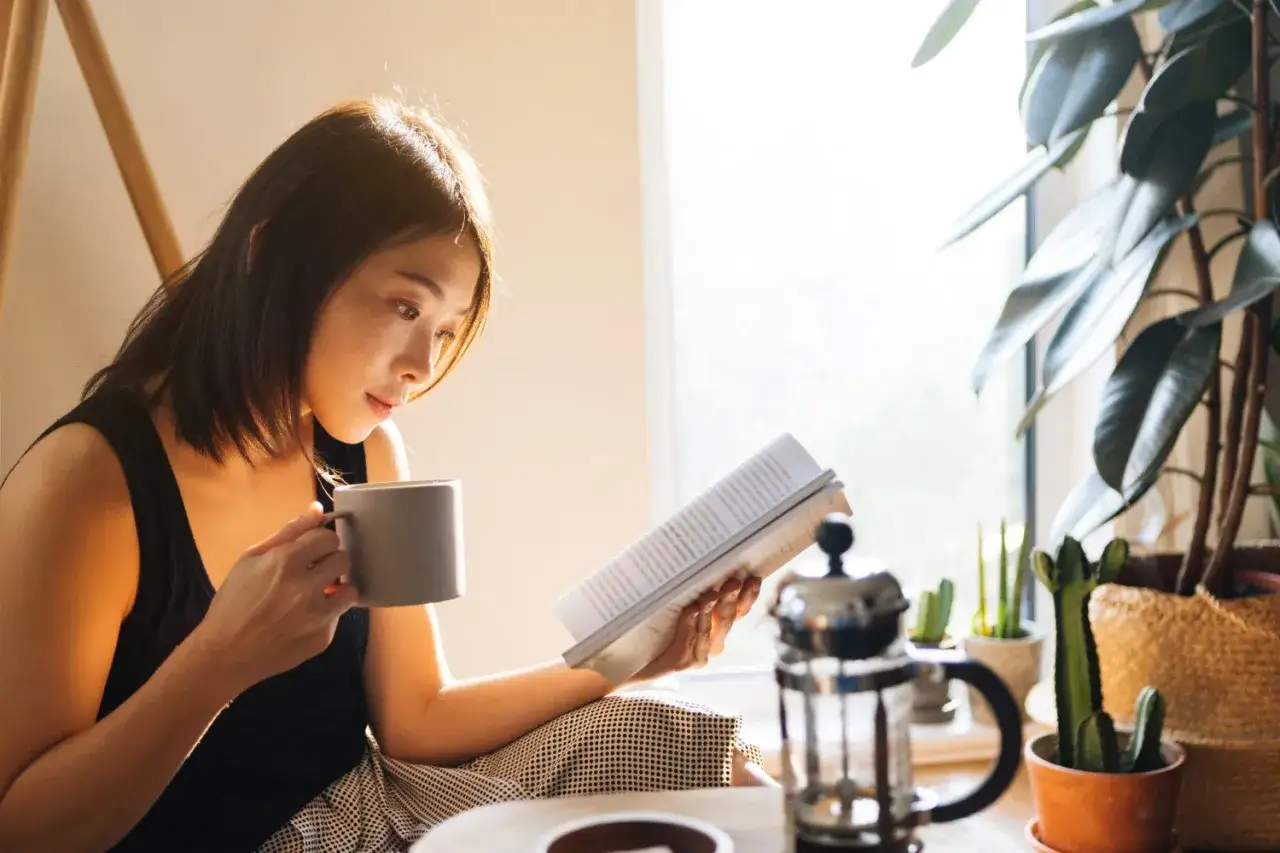 Mujer asi&aacute;tica disfruta de su rutina diaria perfecta, leyendo un libro y tomando caf&eacute; junto a la ventana, rodeada de plantas.
