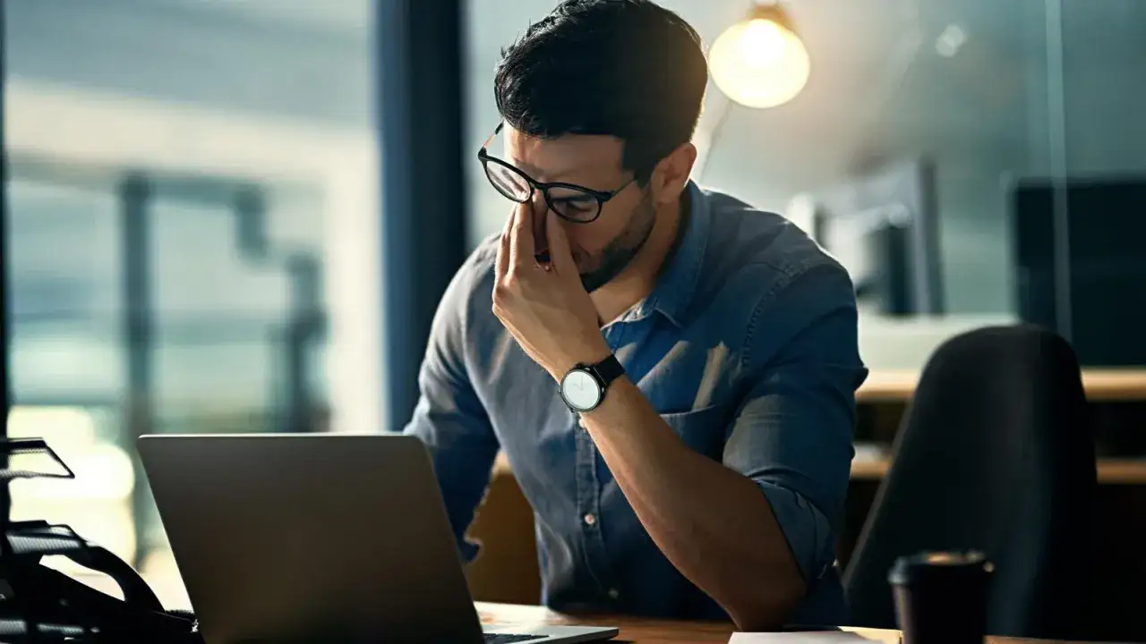 Hombre con gafas se frota los ojos, agotado frente a su port&aacute;til. Un ejemplo de la dificultad en la gesti&oacute;n del estr&eacute;s laboral.
