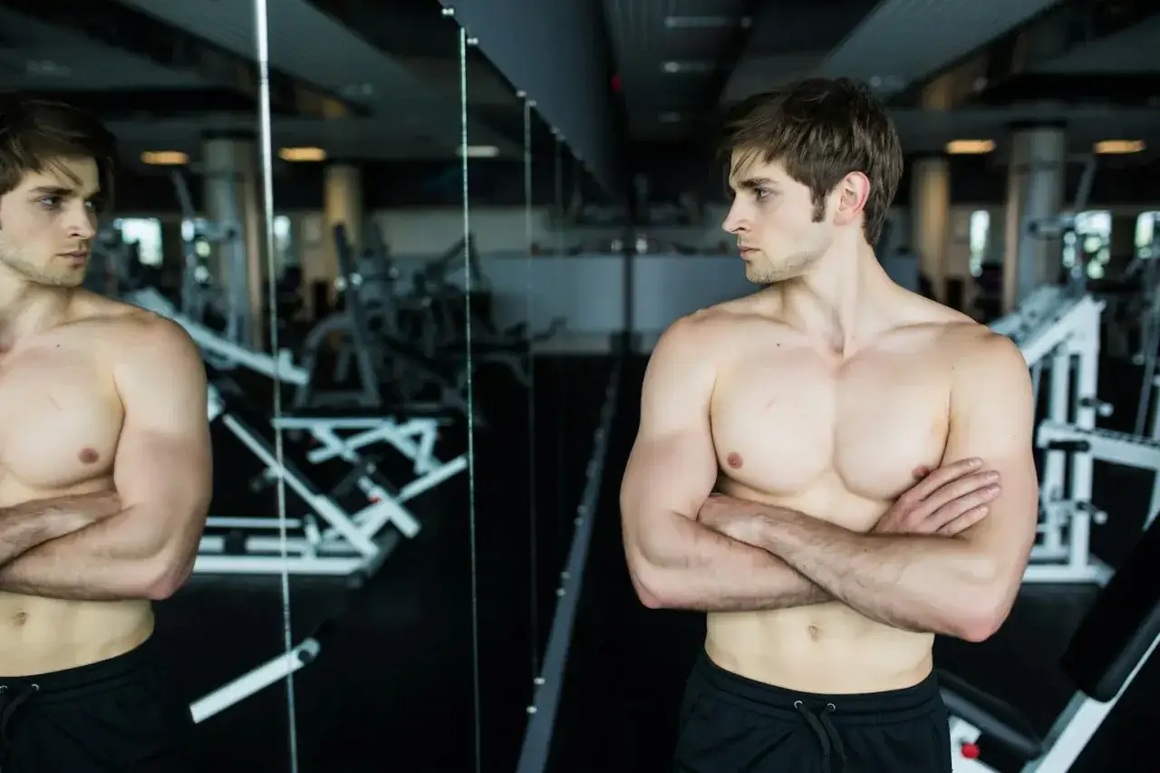 Joven musculoso se mira al espejo en el gimnasio, reflexionando sobre qu&eacute; es la vigorexia y la obsesi&oacute;n por el cuerpo perfecto.