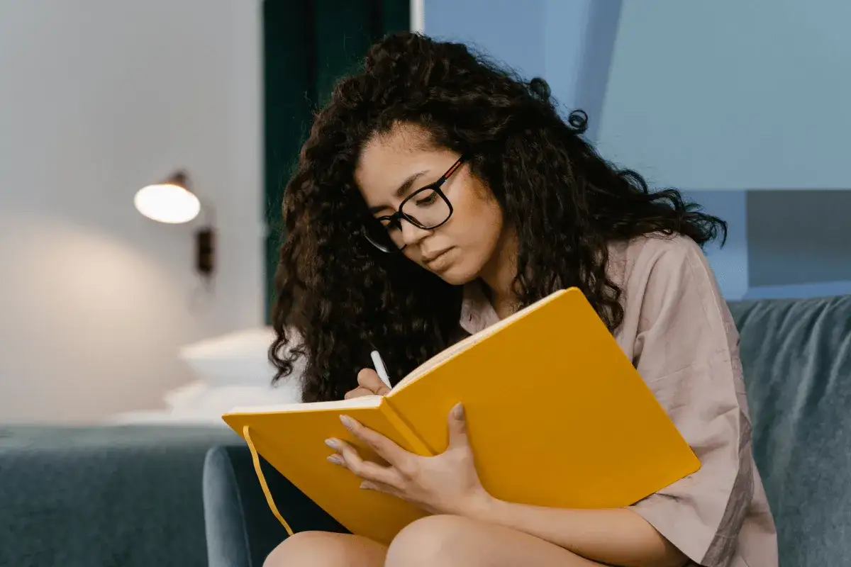 Mujer con gafas escribe en un cuaderno amarillo, buscando mejorar su baja autoestima a trav&eacute;s de la reflexi&oacute;n.