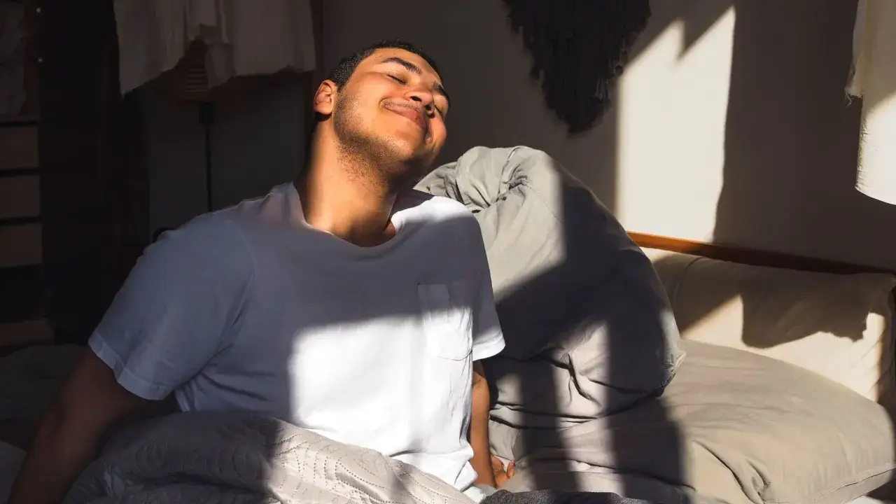 Joven sonr&iacute;e al levantarse a las 5 de la ma&ntilde;ana, sintiendo los primeros rayos de sol en su rostro mientras se estira en la cama.