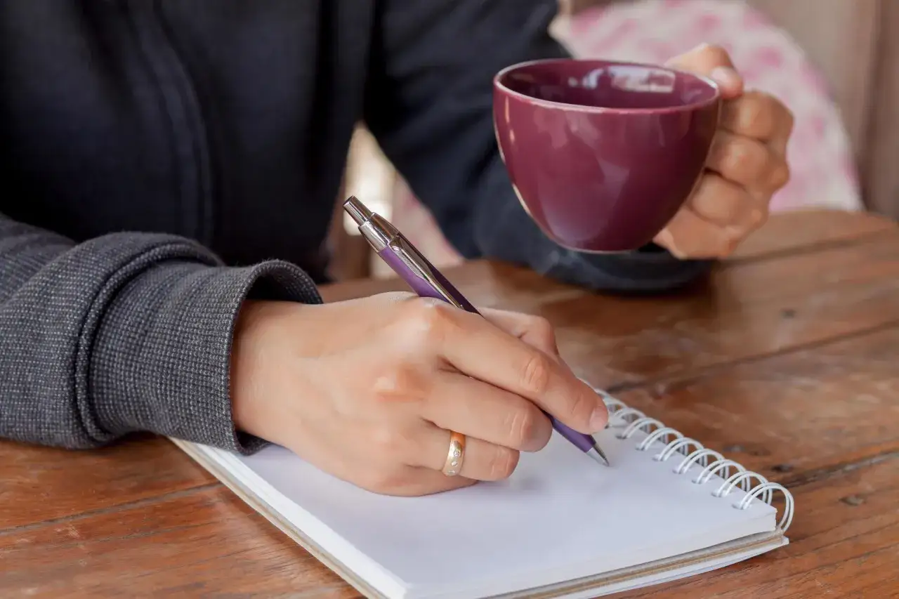 Persona escribe en un cuaderno mientras sostiene una taza de caf&eacute;. Es un momento de reflexi&oacute;n, una forma de practicar que es journaling.