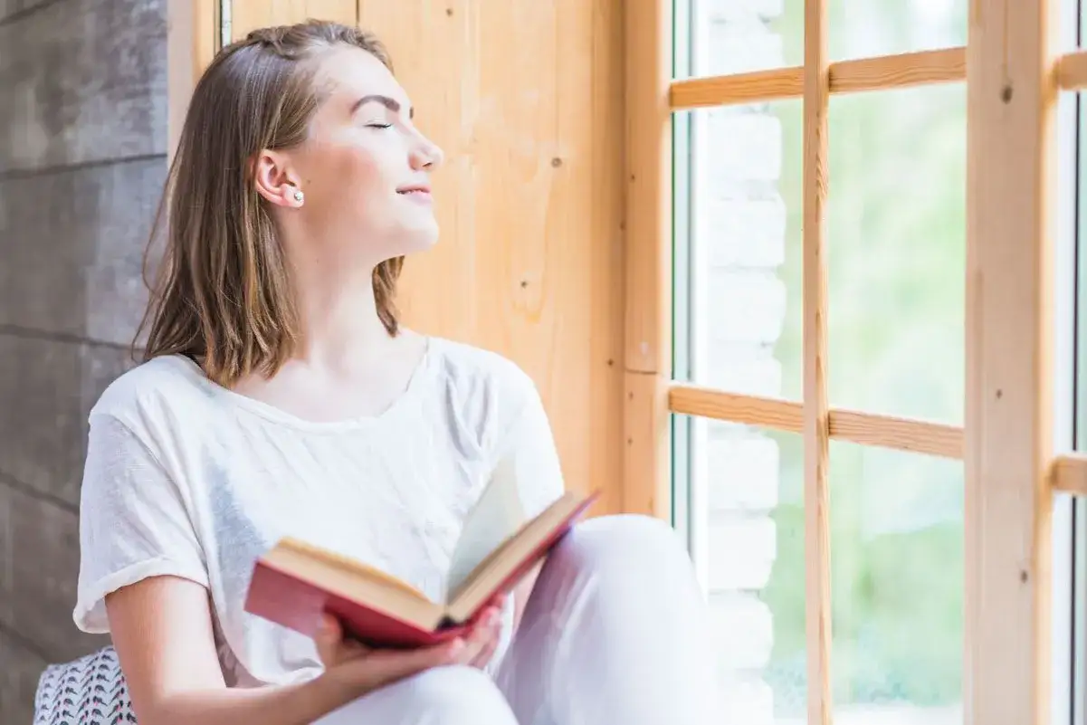 Joven leyendo un libro junto a la ventana, un momento de paz para saber como desengancharse del m&oacute;vil y conectar con uno mismo.