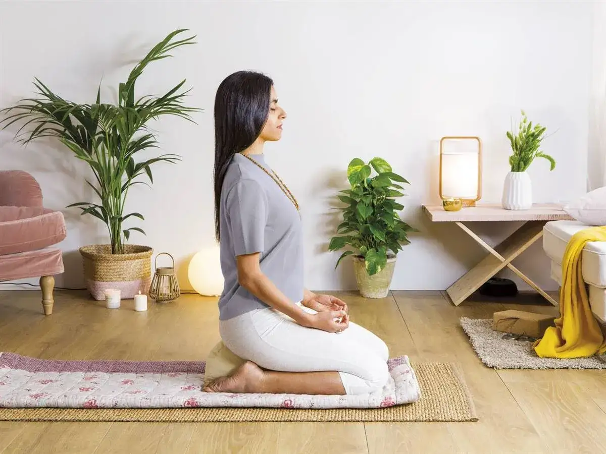 Mujer practicando mindfulness ejercicios de meditaci&oacute;n sentada en un coj&iacute;n, rodeada de plantas y luz tenue.