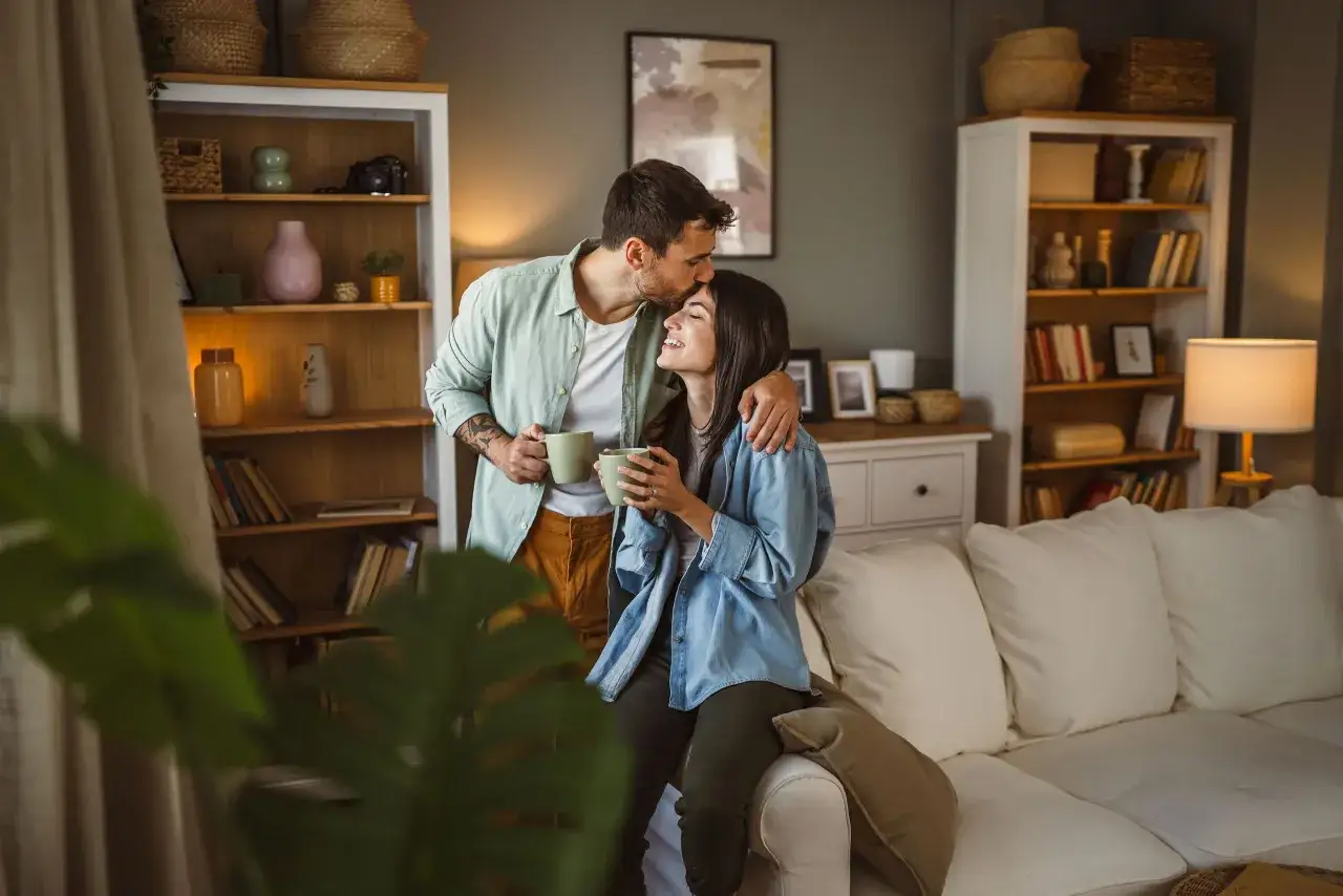 Pareja compartiendo un momento &iacute;ntimo, &eacute;l besa la frente de ella. Su conexi&oacute;n irradia apego seguro, mientras disfrutan de una bebida caliente en su acogedor hogar.