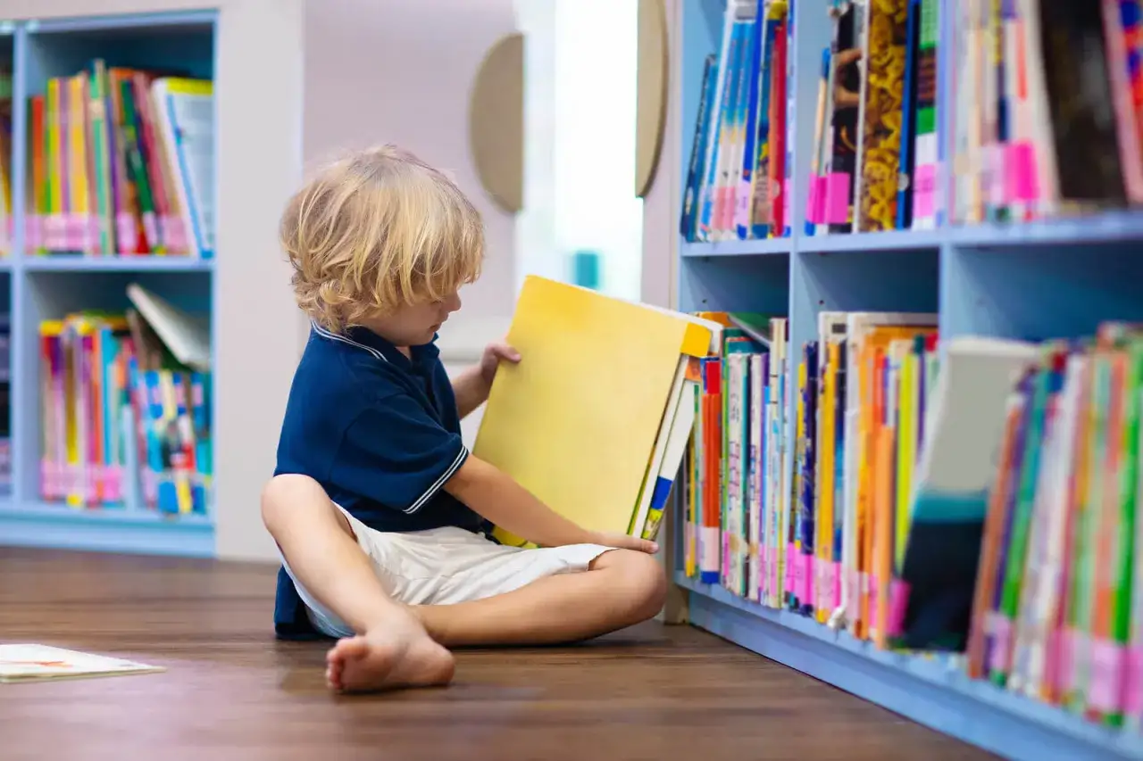 Ni&ntilde;o rubio sentado en el suelo de una biblioteca, eligiendo un libro de una estanter&iacute;a. Ideal para lectura para ni&ntilde;os de 10 a 12 a&ntilde;os.