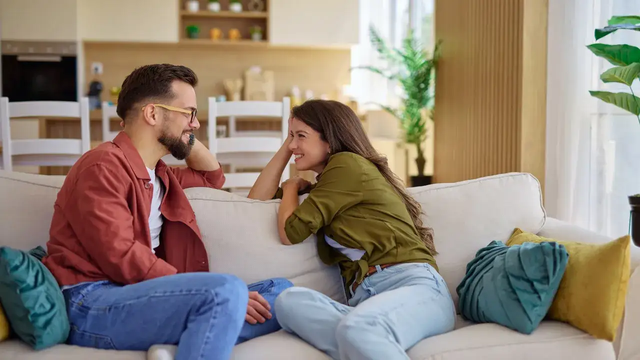 Pareja riendo en un sof&aacute;. &Eacute;l, un hombre de alto valor con barba y gafas, le cuenta algo divertido a ella, que lo mira con complicidad.