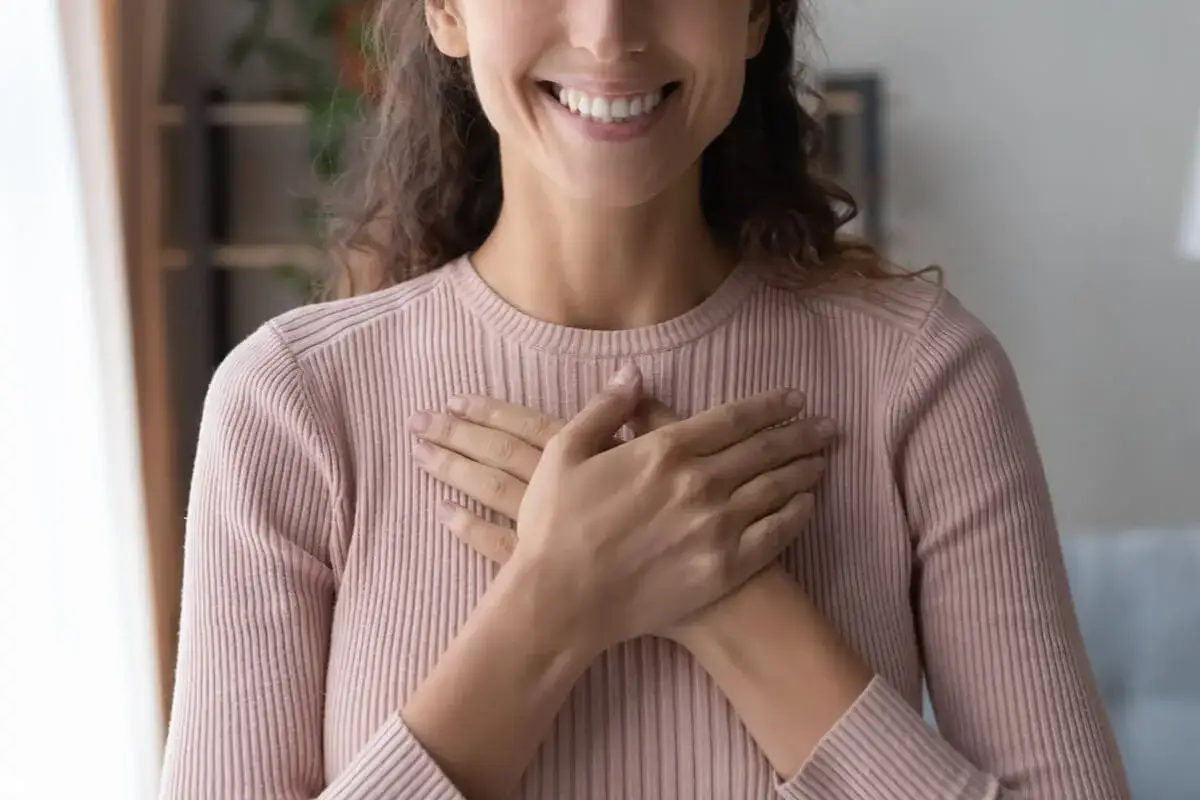 Sonrisa radiante y manos cruzadas sobre el pecho, un gesto de profundo amor propio y gratitud.