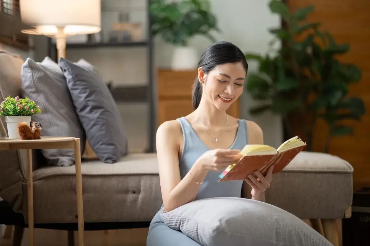 Mujer sonr&iacute;e leyendo libros para subir el &aacute;nimo, sentada en el suelo con cojines, rodeada de un ambiente acogedor.