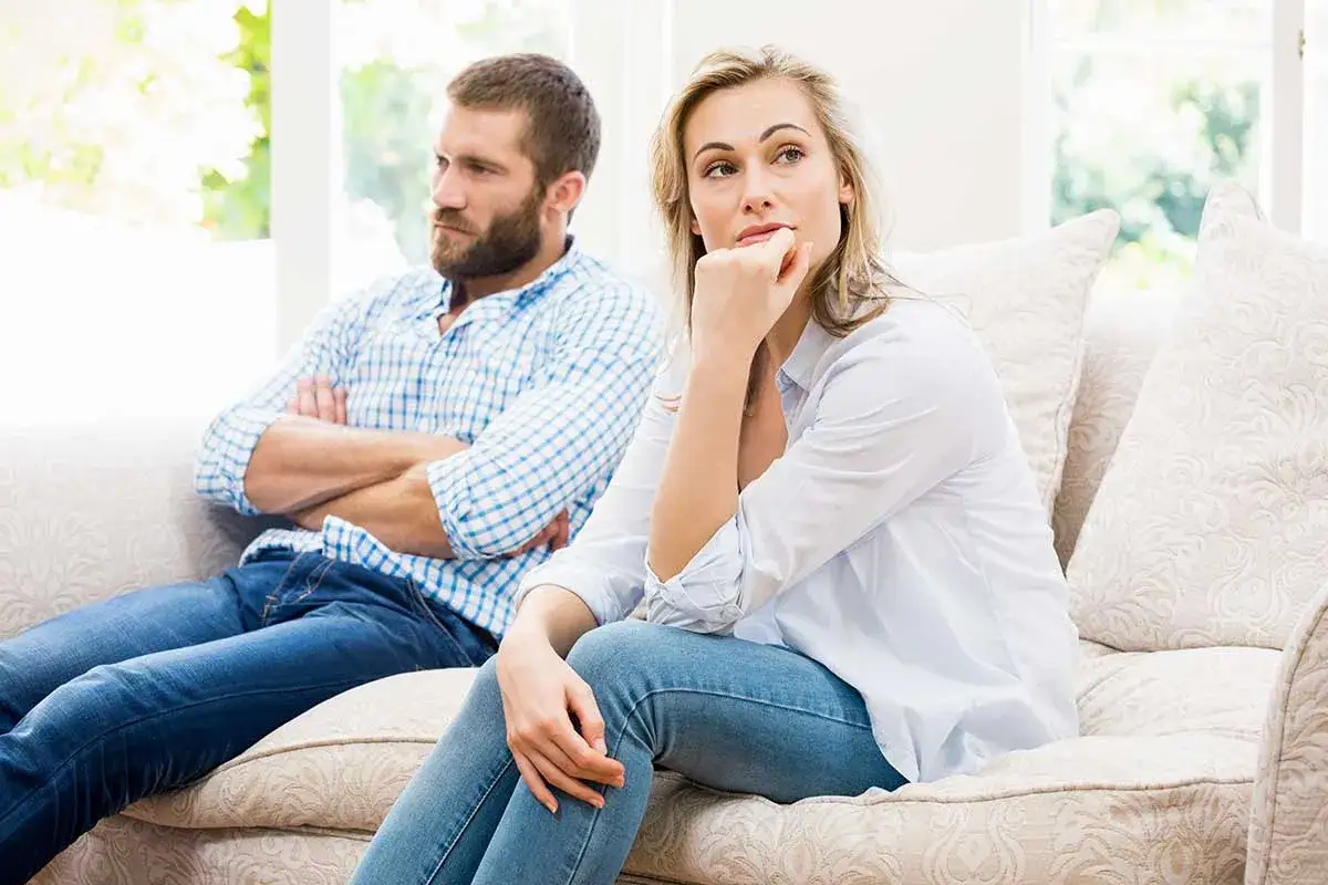 Pareja sentada en un sofá, él con los brazos cruzados y mirada perdida, ella pensativa. Quizás reviviendo celos retrospectivos.