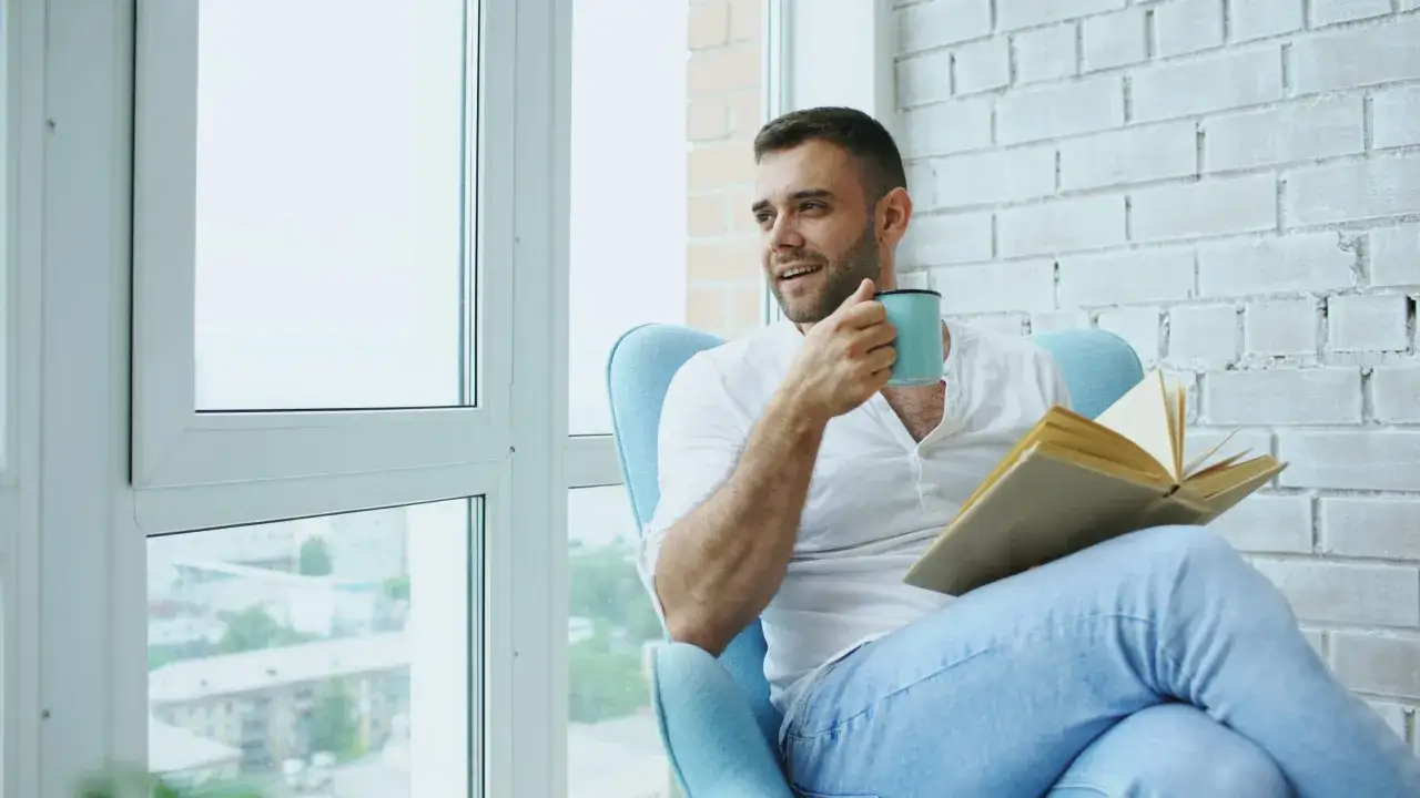 Hombre joven con taza y libro abierto, disfrutando de un momento tranquilo. Ideal para encontrar nuevos libros para leer en ingl&eacute;s.