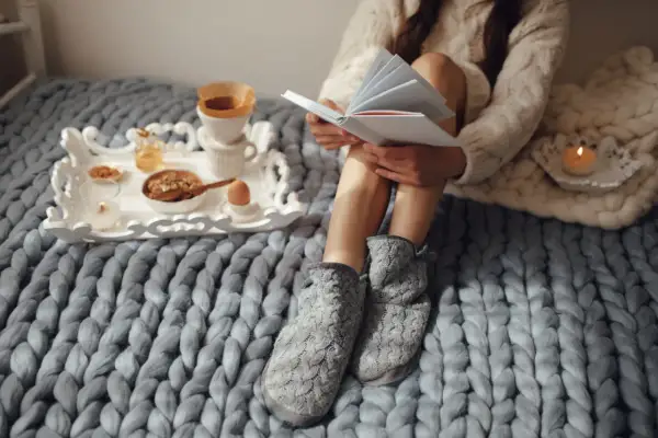 Mujer lee historias basadas en hechos reales mientras desayuna en la cama, envuelta en mantas y con zapatillas.