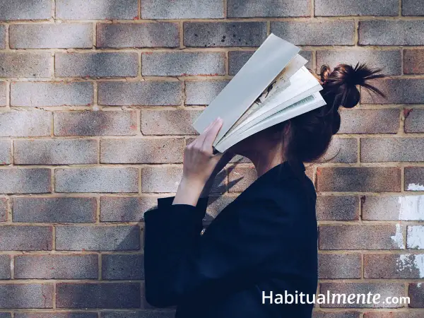 Una persona se esconde detrás de un libro abierto, sumergida en sus hábitos de lectura. La luz y la sombra juegan sobre el ladrillo.