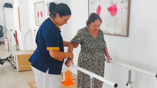 Terapeuta ayuda a anciana a caminar con barras paralelas, un paso importante en varias áreas de la vida.