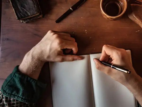 Manos escribiendo en un cuaderno abierto, buscando inspiración para frases bonitas para dedicar un libro. Una pluma, un libro antiguo y una taza de café completan la escena.