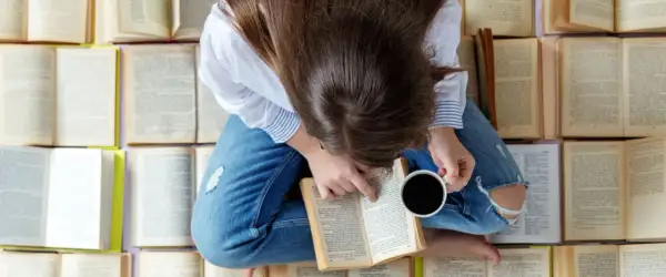 Una persona rodeada de libros que hay que leer antes de morir, con una taza de café en la mano, sumergida en la lectura.