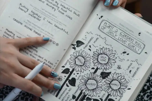 Un diario abierto muestra dibujos de girasoles y la frase "PLANT A GARDEN FULL OF SELF LOVE, GIRL!". Es un ejemplo de que es journaling, una práctica para la autoexpresión.