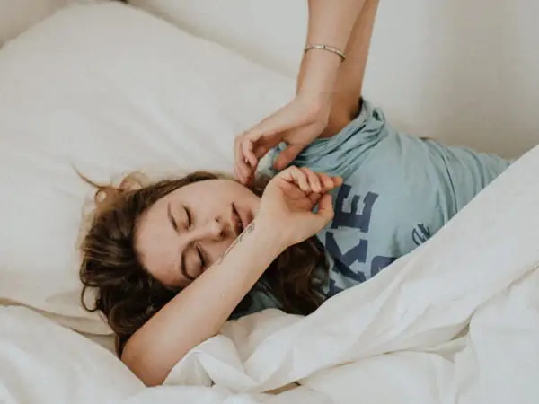 Una mujer se despierta en la cama, con los ojos cerrados y una expresión de agotamiento. Se pregunta porque está tan cansada y solo quiere dormir.