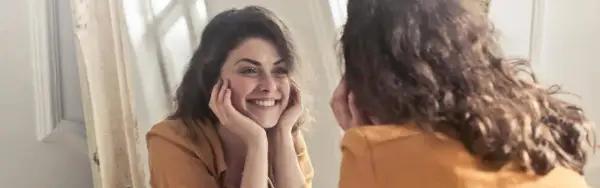 Una mujer sonríe a su reflejo, preguntándose "quien soy" mientras se mira en el espejo.