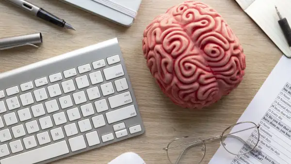 Un cerebro de juguete, teclado, bolígrafo y gafas sobre un escritorio, sugiriendo el modelo biopsicosocial.