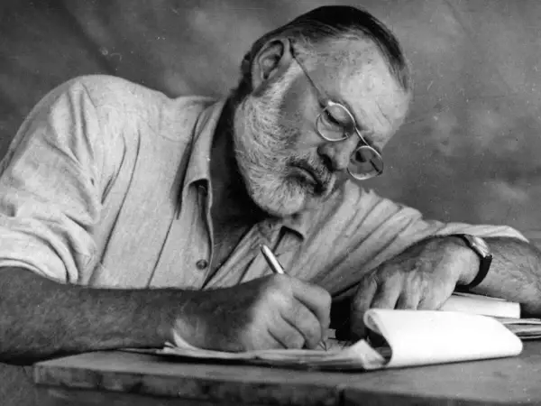 Ernest Hemingway, con gafas y barba, escribe concentrado. Sus **ernest hemingway frases** nacen de esta dedicación.