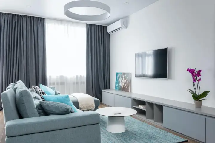 Sofá gris azulado con cojines turquesas y blancos, mesa de centro blanca, alfombra a cuadros, mueble de TV gris y orquídea morada. Un espacio que inspira una vida minimalista.